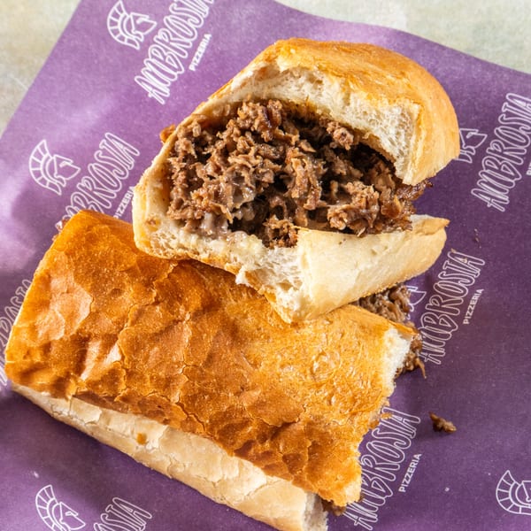 A sliced cheesesteak sandwich with melted cheese and steak on a purple Ambrosia Pizzeria wrapper.