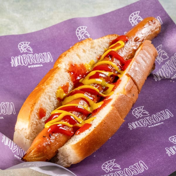 A hot dog with ketchup and mustard on a bun, resting on purple Ambrosia Pizzeria branded paper.