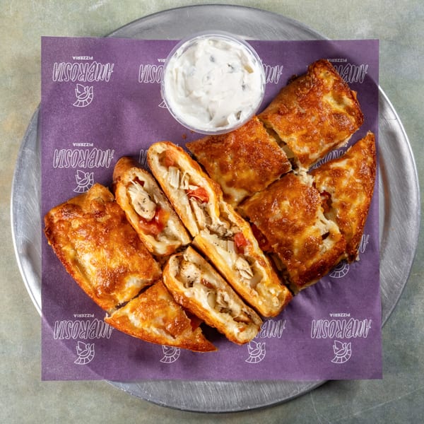 Ambrosia Pizzeria's golden-brown calzone, sliced to reveal chicken and tomatoes, with a side of dipping sauce.