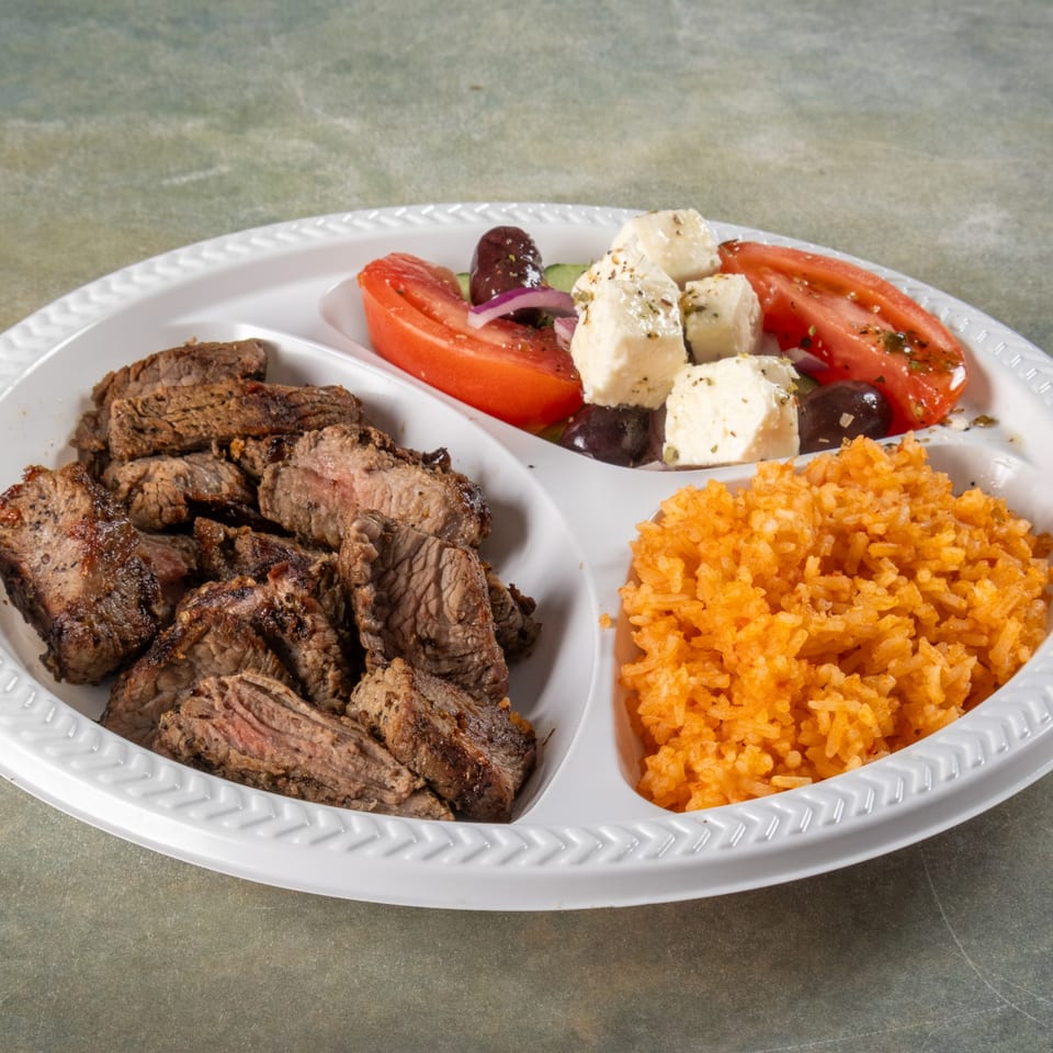 A divided plate with grilled steak, orange rice, and a Greek salad with tomatoes, olives, and feta.